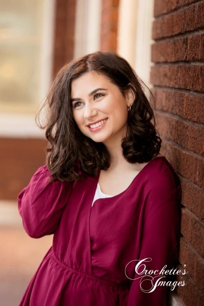 Senior girl photo in maroon dress leaning on a brick wall with amazing smile