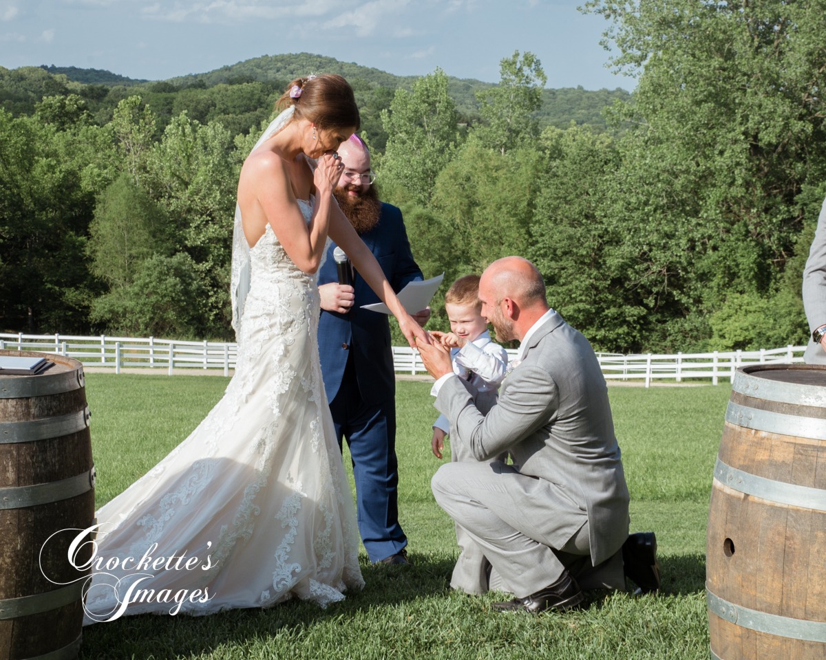 Dad and son put rings on mom's finger at an outdoor wedding ceremony