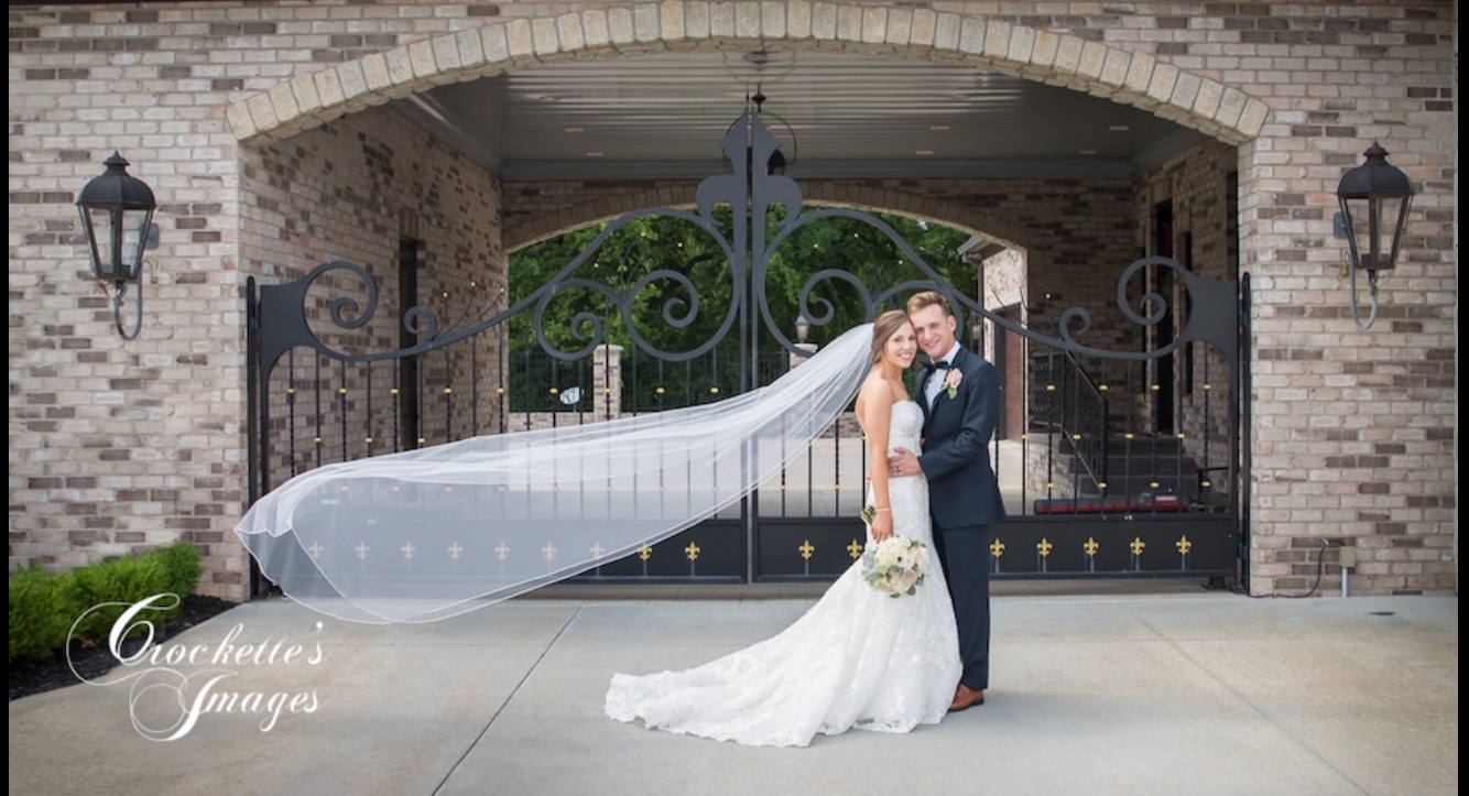 Elegant wedding photo of a couple infront of a fancy metal gate with long veil blowing in the wind