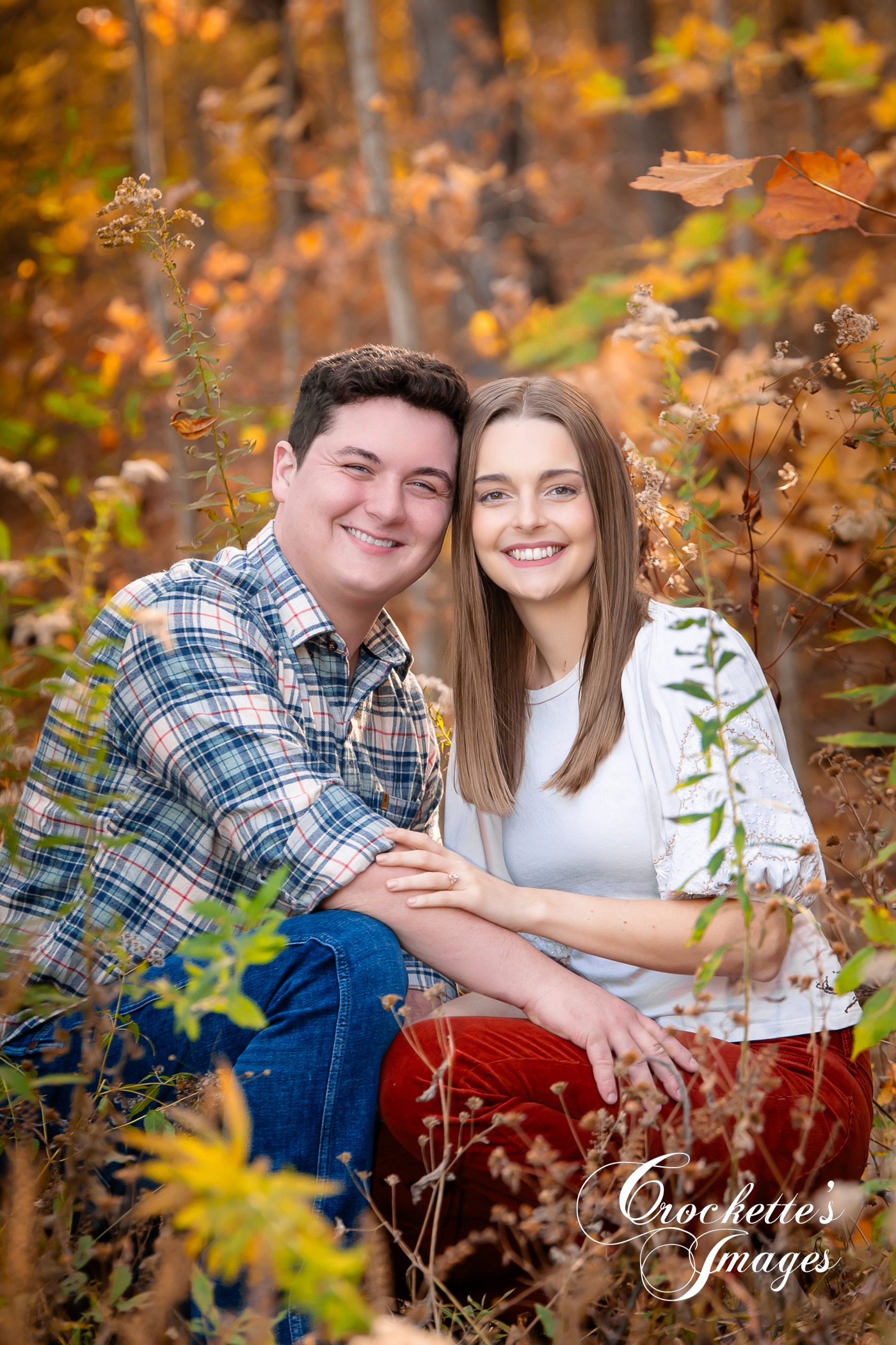 Fall engagement photo with couple sitting in fall weeds with woods in the background