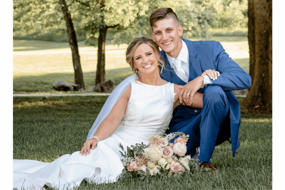 Bride and groom pose in the green grass under the trees in the summer shade following their wedding ceremony with the bride's dress neatly laid beside her.