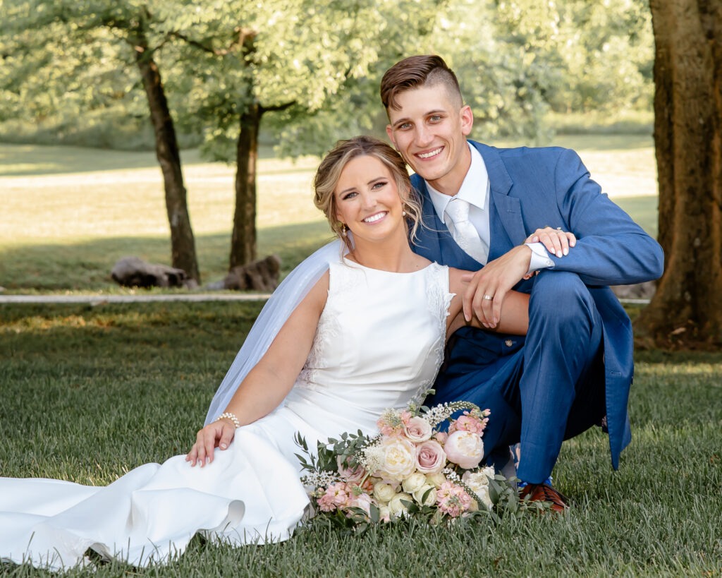 Bride and groom seated in the grass beneath shade trees, posing for a wedding portrait with the bride’s dress spread out to the side.