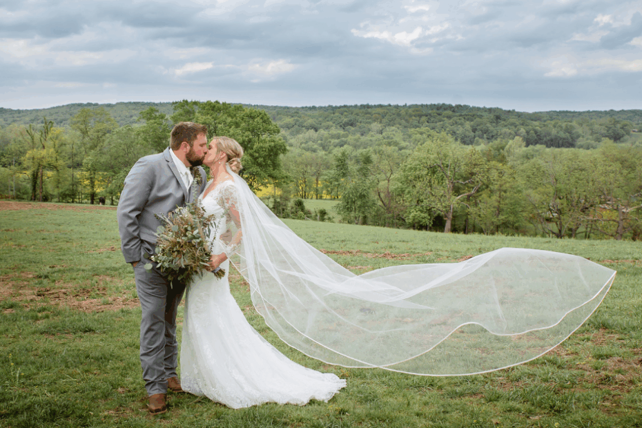 Bride and groom kiss in a field on the farm with the bride's veil blowing behind her, with the deep colors of summer green surrounding them.