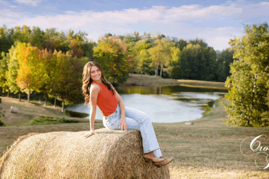 Senior girl posing for her senior photos during late summer in a farm setting on a round hay bale.