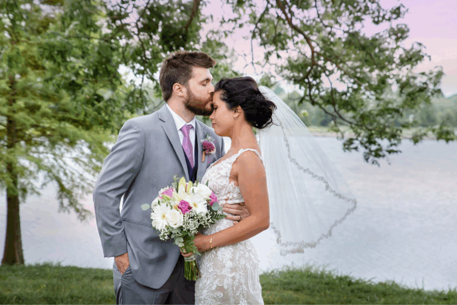 Groom kisses the head of his new bride as her veil blows in the wind behind her at sunset.