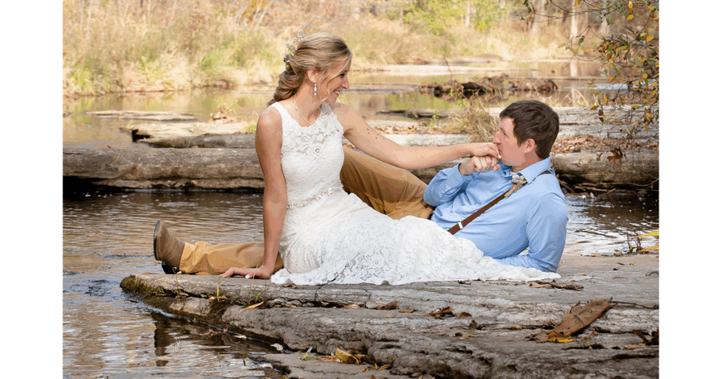 Bride and groom lie on rocks in a creek as groom lovingly kisses his bride's hand. 