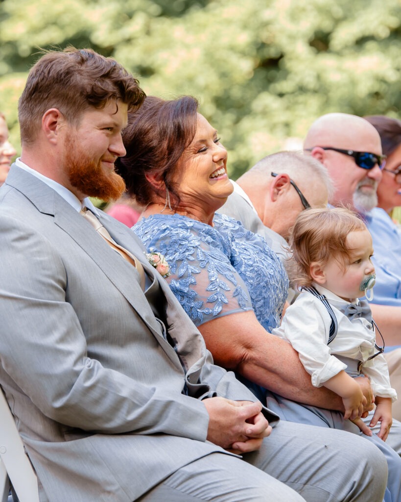 Smiling wedding guests watch as the bride and groom exchange vows during an outdoor wedding ceremony.